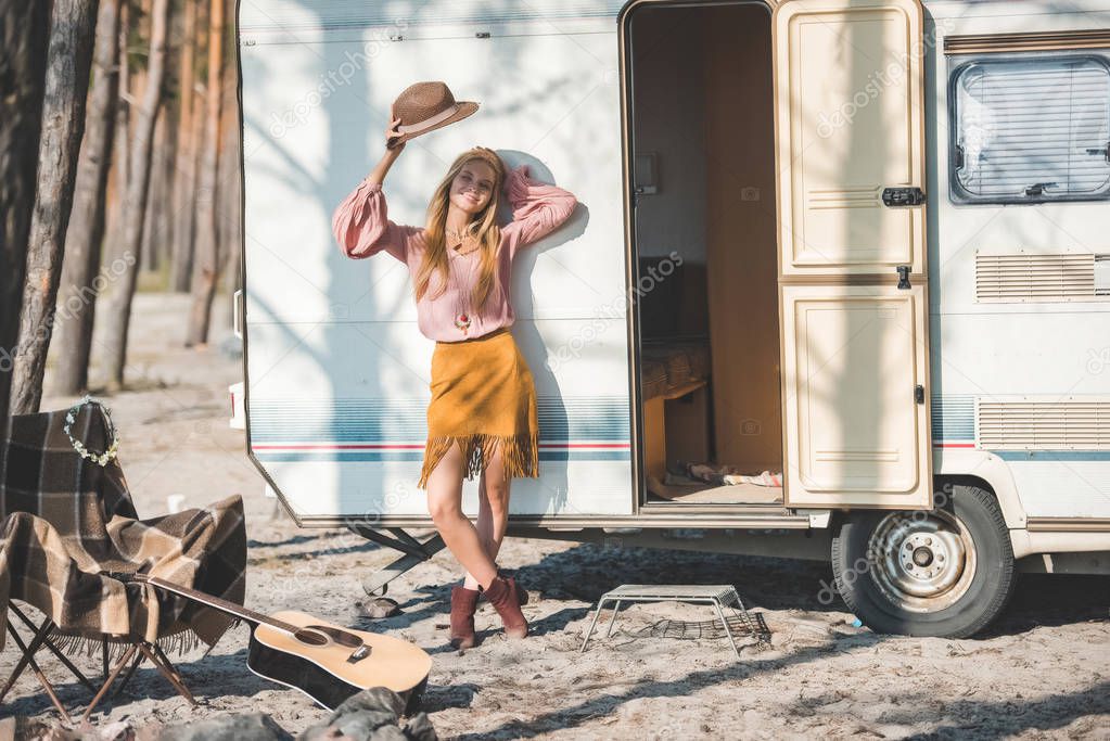 young hippie woman with hat posing near trailer with guitar in forest