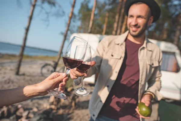 young man and woman clinking with glasses of wine walking near campervan