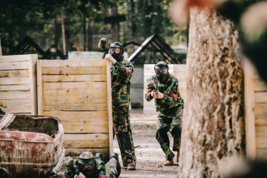 selective focus of paintball team in uniform and protective masks playing paintball with marker guns outdoors clipart