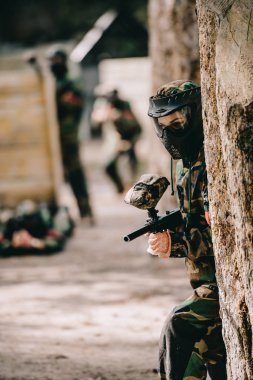 selective focus of female paintball player in goggle mask and camouflage aiming by paintball gun near tree outdoors  clipart