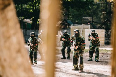 selective focus of paintball team in uniform and protective masks playing paintball with marker guns outdoors clipart