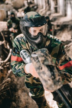 selective focus of male paintball player in goggle mask and camouflage aiming by paintball gun from ditch outdoors clipart