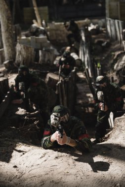 paintball team in camouflage and protective masks aiming by marker guns from ditch outdoors  clipart