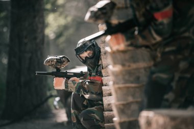 selective focus of paintball player in protective mask holding marker gun and his teammate hiding behind wooden wall outdoors clipart