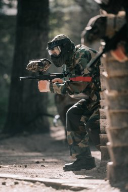 selective focus of male paintball player in goggle mask and camouflage aiming by paintball gun outdoors  clipart