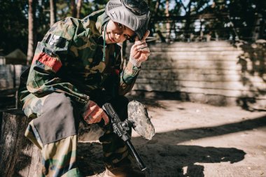Genç kadın paintballer kamuflaj üniforma ile paintball silahı açık havada oturma içinde gülümseyen 