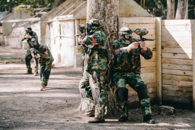 male paintball players in camouflage and protective masks hiding behind tree and shooting by marker guns outdoors  clipart