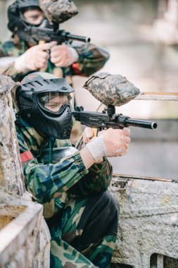 side view of concentrated paintball player in protective mask aiming with marker gun outdoors clipart