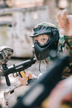 male paintballer in goggle mask and camouflage holding marker gun and pointing by finger outdoors  clipart