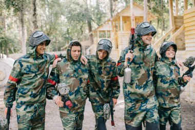 Paintball oyuncular kamuflaj açık havada birbirlerine kucaklayan marker silahlarla gülüyor. 