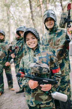 beautiful happy female paintballer in uniform holding paintball gun while her team standing behind outdoors  clipart