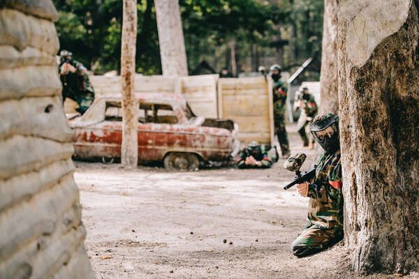 female paintball player in goggle mask and camouflage sitting with paintball gun while her team sitting behind near broken car outdoors 