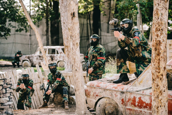 paintball team in uniform and protective masks playing paintball with marker guns on broken car outdoors 
