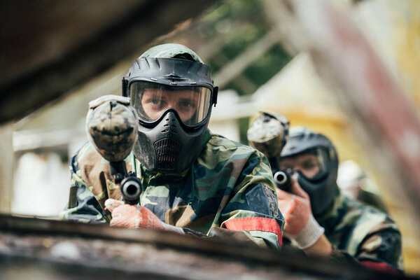 male paintballer and his team in uniform and protective masks aiming by paintball guns outdoors