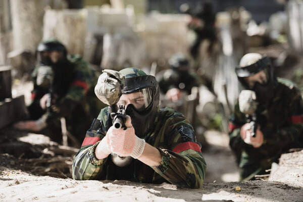 selective focus of concentrated paintball player in protective mask aiming with marker gun and his team on background outdoors