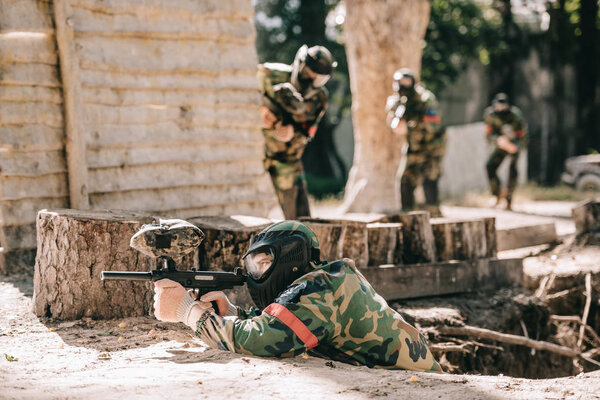 selective focus of paintball player laying on ground and aiming with marker gun and his team hiding behind wooden wall outdoors