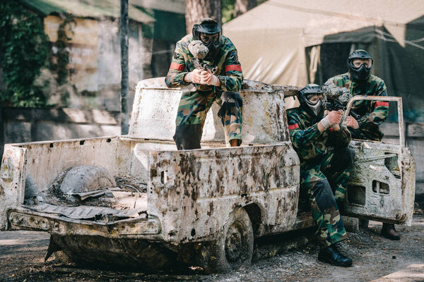 пейнтбольная команда в форме и защитных масках, нацеленных на пейнтбольные пистолеты из сломанной машины на открытом воздухе
 