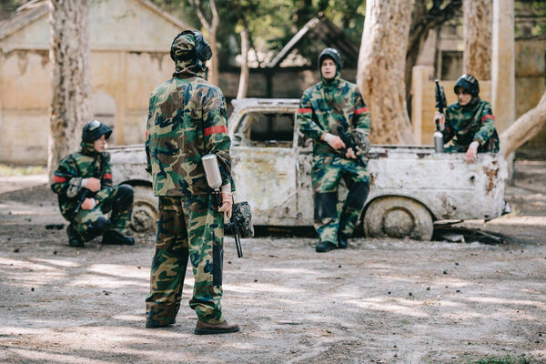 paintball team in camouflage uniform with marker guns resting near broken car outdoors 