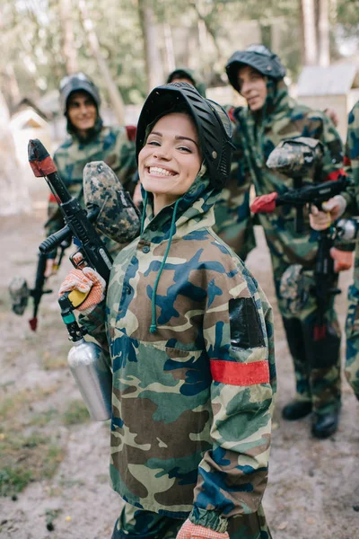 屋外の後ろに彼女のチームに立って中ペイント ボール銃を保持している制服で魅力的な笑顔の女性 Paintballer — ストック写真