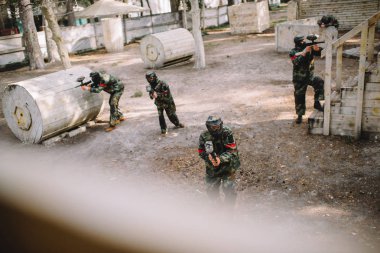 high angle view of paintball team in uniform and protective masks playing paintball with markers guns outdoors clipart