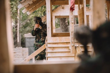 selective focus of male paintball player in goggle mask and camouflage aiming by paintball gun on wooden tower clipart
