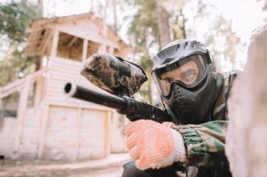serious male paintball player in goggle mask and camouflage aiming by paintball gun outdoors  clipart