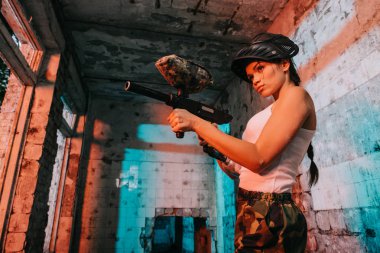 low angle view of young female paintballer in camouflage and white singlet aiming by marker gun in abandoned building