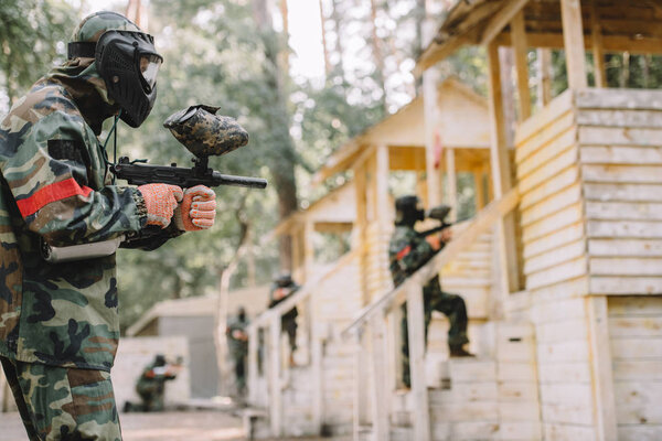 side view of paintballer in camouflage and protective mask aiming by marker gun while his team standing behind outdoors 