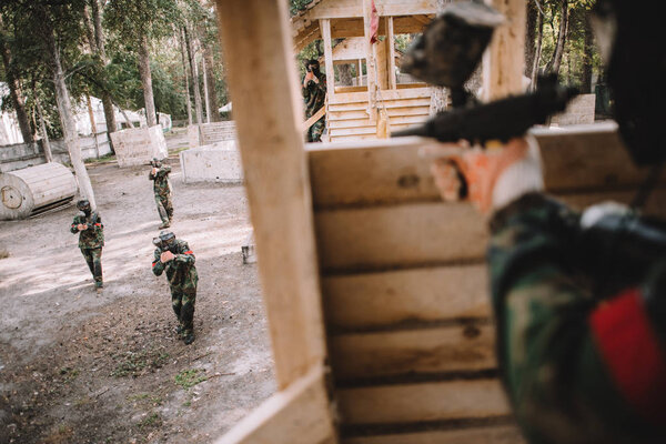 cropped image of paintball player in camouflage uniform aiming by paintball gun from wooden tower outdoors 