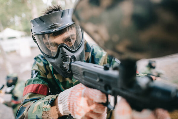 selective focus of male paintball player in goggle mask and camouflage aiming by paintball gun outdoors 