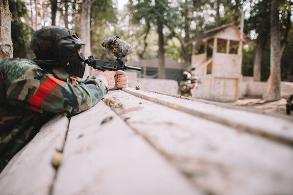 selective focus of paintball player in protective mask and camouflage aiming by marker gun outdoors 