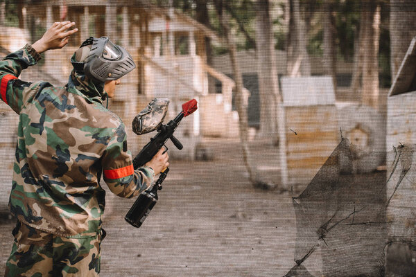 rear view of male paintball player in goggle mask and camouflage with paintball gun standing near net outdoors 