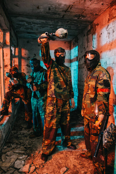 male paintball player aiming by gun at camera while his paintball team in uniform and protective masks standing near in abandoned building