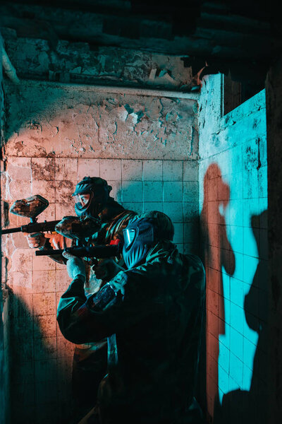 side view of paintball team in uniform and protective masks playing paintball with marker guns in abandoned building