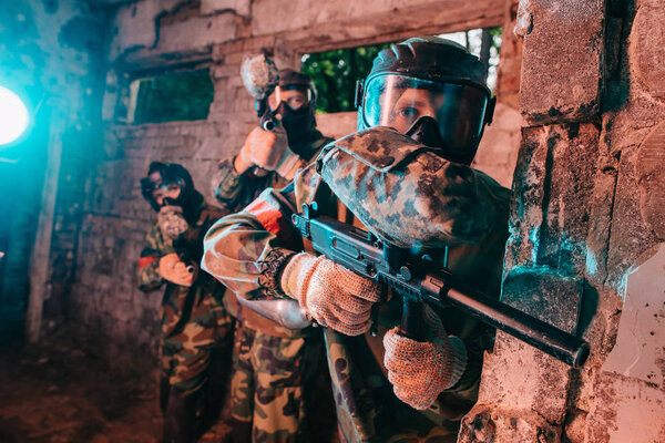 selective focus of paintball team in uniform and protective masks playing paintball with marker guns in abandoned building