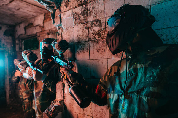 male paintball player in goggle mask and camouflage uniform hiding behind wall and aiming marker gun at player of other team in abandoned building 