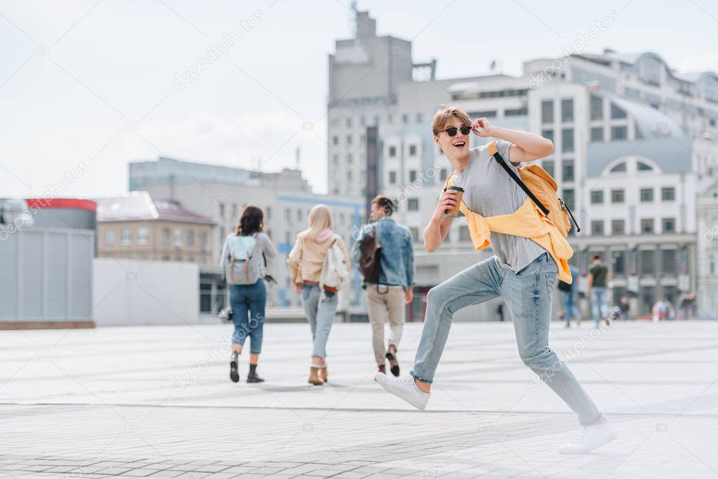 excited stylish traveler with coffee to go and backpack having fun on street with friends behind