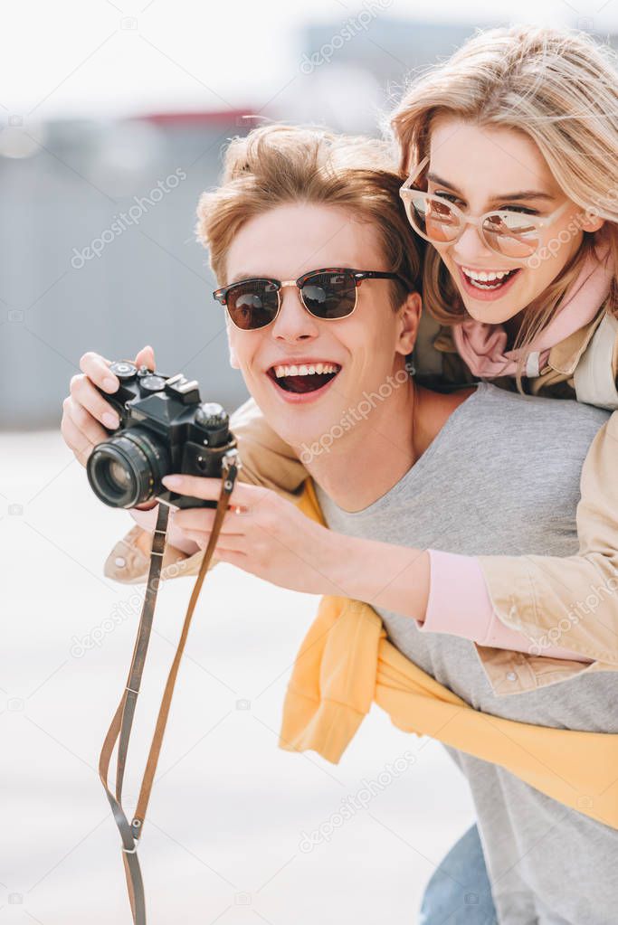 smiling tourist giving piggyback to happy girlfriend while she taking photo on camera
