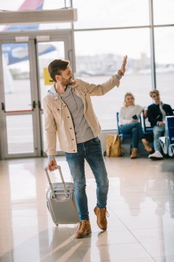 mutlu genç adam el sallayarak ve uzağa bakarak havaalanında bavul ile