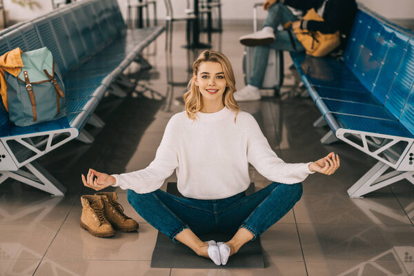 девушка медитирует в позе лотоса и улыбается в камеру, ожидая в терминале аэропорта
