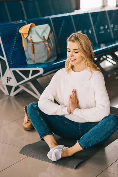улыбающаяся молодая женщина медитирует в позе лотоса, ожидая полета в терминале аэропорта
