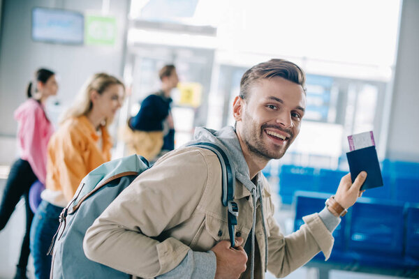 happy young man with backpack holding passport with boarding pass and smiling at camera in airport