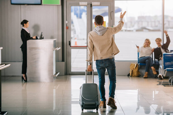back view of young man with suitcase waving hand to friends in airport