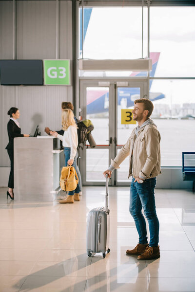 вид сбоку улыбающегося молодого человека, стоящего с чемоданом в аэропорту
