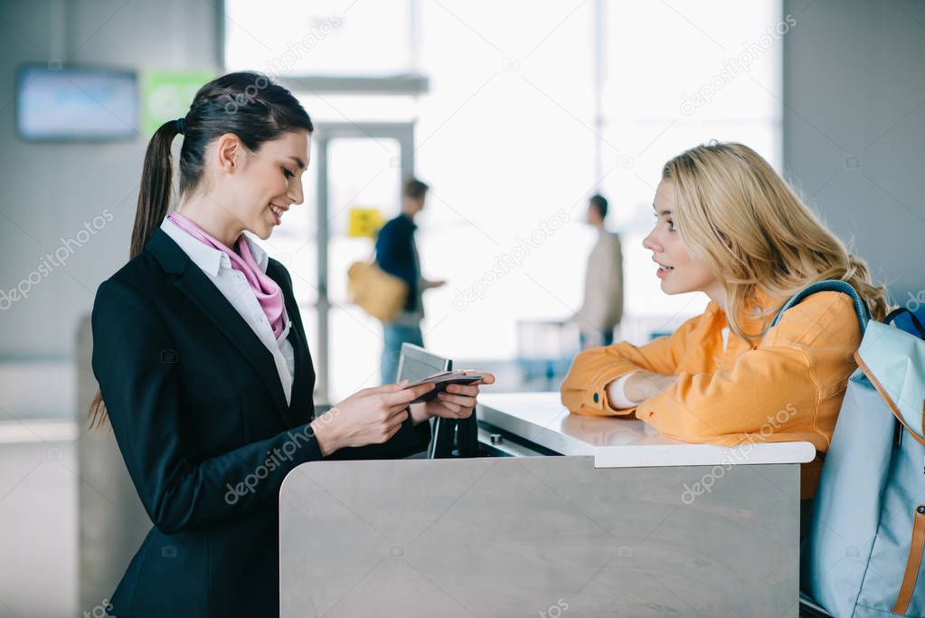 side view of smiling airport worker checking documents of young female traveler at check-in desk  