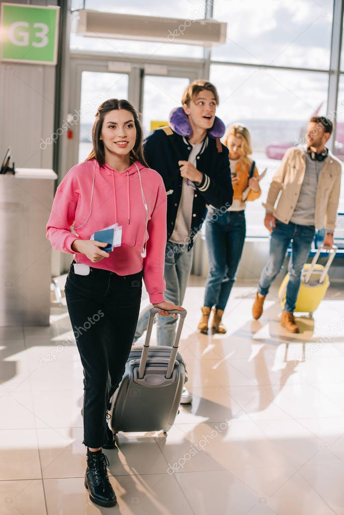 smiling young woman holding suitcase, passport and boarding pass while traveling with friends