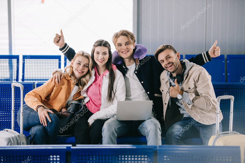 happy young friends smiling at camera and showing thumbs up while waiting for flight in airport terminal 