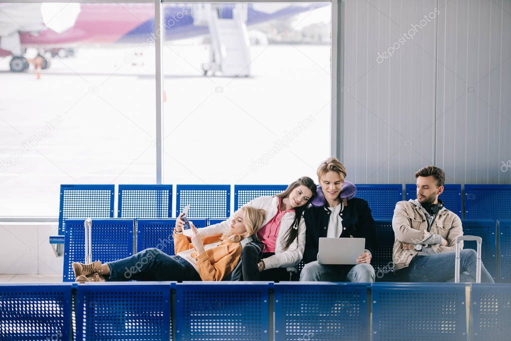 young friends holding documents and using laptop while waiting for flight in airport terminal