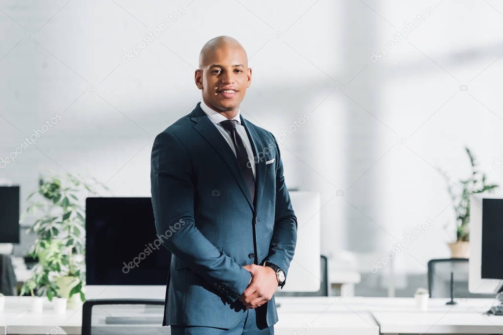 confident young african american businessman standing in office and smiling at camera