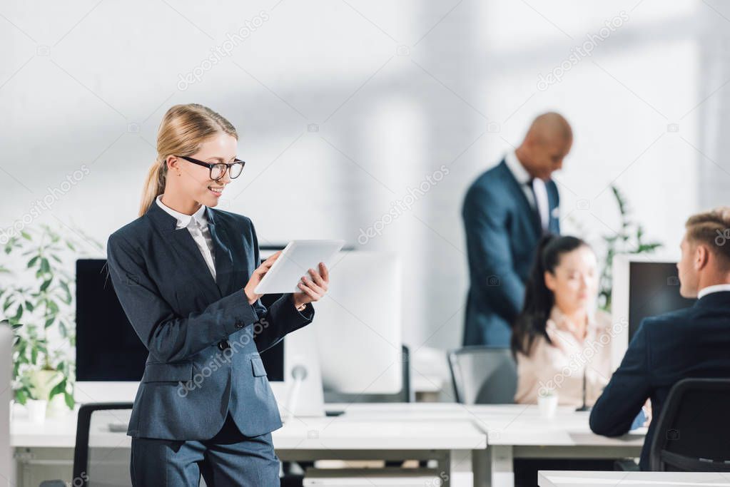 smiling young businesswoman in eyeglasses using digital tablet and colleagues working behind in open space office